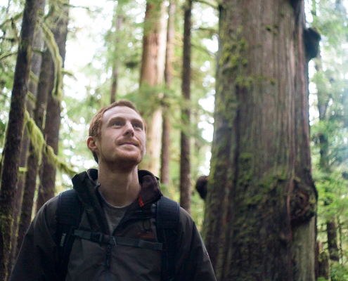 TJ stands in the forefront wearing a black hoodie. Behind him stands a number of old-growth cedars and other ancient trees.