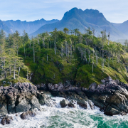 A turquoise ocean splashes against craggy rocks with lush, green old-growth forest and blue-hued mountains in the background.